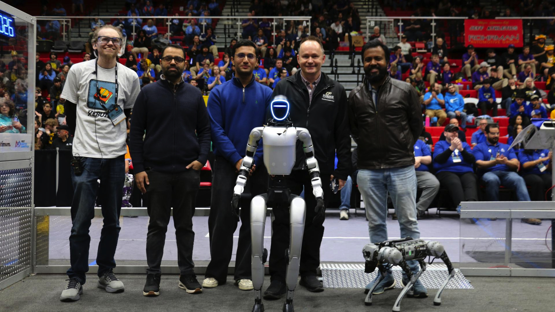 A group of five men stand on a robotics competition field alongside two robots—a humanoid robot in the center and a smaller quadruped robot to the right—while a crowd of spectators fills the bleachers in the background.