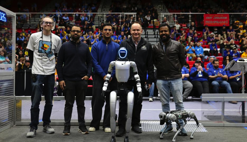 A group of five men stand on a robotics competition field alongside two robots—a humanoid robot in the center and a smaller quadruped robot to the right—while a crowd of spectators fills the bleachers in the background.